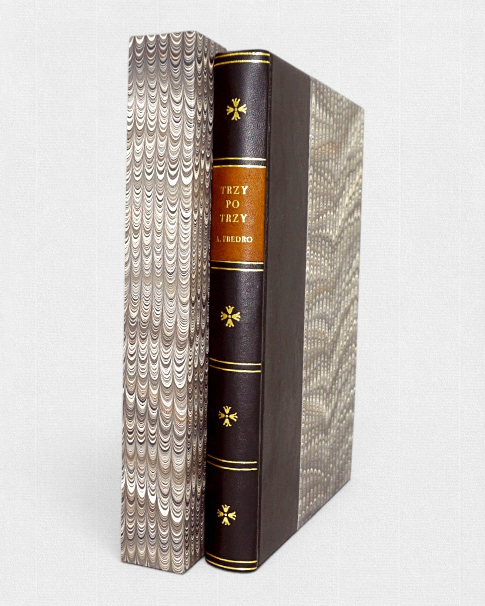 Książka „Trzy po trzy” Aleksandra Fredry w półskórkowej oprawie ze złoceniami, obok marmurkowego etui ochronnego