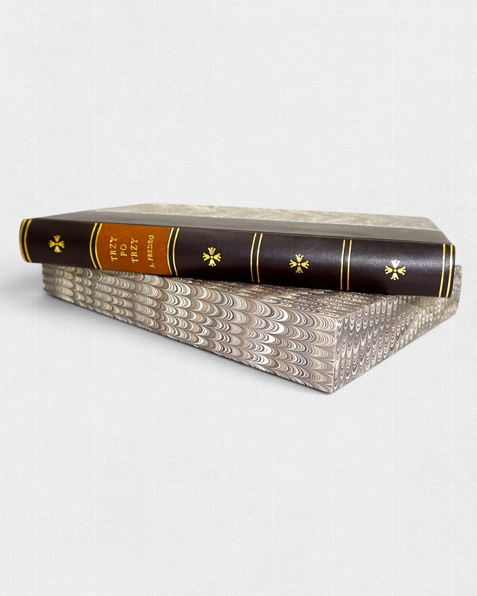 Książka „Trzy po trzy” Aleksandra Fredry w półskórkowej oprawie ze złoceniami, ułożona na marmurkowym etui, widok z boku